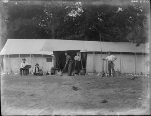 Five unidentified young men in hats with wood burner stove and pot, accordion, broom, dog and axe, in front of two tents with awning and 'Myrtle Camp' sign, trees beyond, [Sumner?], Christchurch