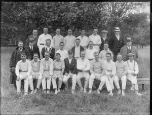 Group portrait of unidentified men [cricket team?] mostly in 'whites' with coaches, one man in blazer and cap with badge, on long grass with lines of trees beyond, probably Christchurch region