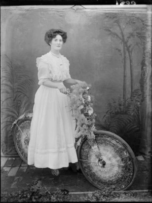 A full length portrait of an unidentified woman sitting on a decorated bicycle, taken in the studio, probably Christchurch district