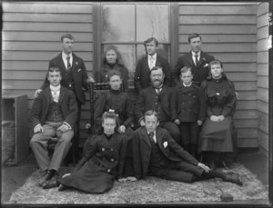 Family group portrait in front of a wooden house, unidentified older parents with their six sons and three daughters on carpet, probably Christchurch region