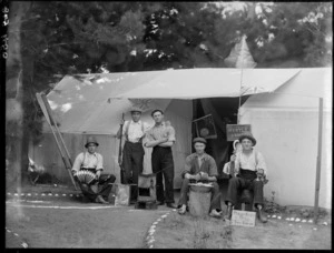 Group of five unidentified men in work clothes, at a campsite, holding various props including a rifle and an accordian, with sign reading 'Myrtle Camp' on tent behind, possibly Christchurch district