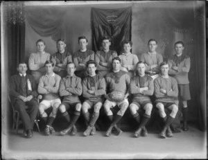 Studio portrait of the Woolston 4th Grade Rugby [league?] Football Team, 1920 winners, unidentified players in uniform with ball and coach wearing a ribbon, Christchurch