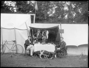 Group dining at 'Ngaio Camp', showing three unidentified young men and a dog at a campsite, sitting at a table covered with a cloth and laid with a vase of flowers, tea service, and food, alongside tents and a bicycle, possibly Christchurch district
