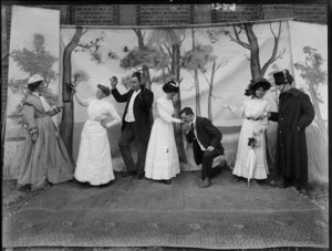Unidentified actors with false backdrop on grass with brick wall behind, older couple remonstrating with 'boot in hand' with maid, man on knee kissing hand of young lady, soldier with gun talking to young woman, probably Christchurch region