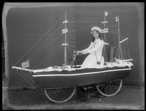 Unidentified girl on a bicycle decorated as a ship Terra Nova, taken outdoors with a dark backdrop, possibly Christchurch district