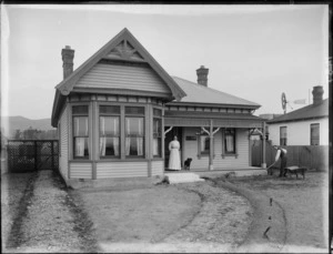 Unidentified woman standing on the porch with a dog, and a unidentified man working in the garden in front of a house named Awahuri, possibly Christchurch district