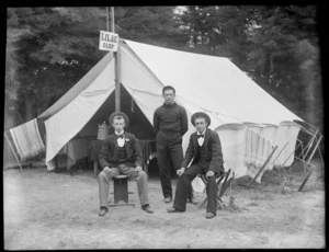 Three unidentified members of the Lilac Camp, two men sitting on wooden stools and one man standing behind them in front of an open tent, showing a table inside the tent with a basket on it, possibly Christchurch district