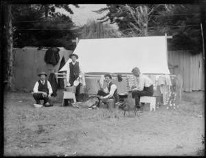 Four unidentified men next to the tent doing odd jobs, showing two men doing the dishes, one man chopping wood and the other man cooking, possibly Christchurch district