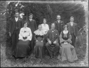 Members of a unidentified family, showing five men and four women, with three women and a man sitting on chairs and four men and a woman standing behind them, in front of macrocarpa tree, possibly Christchurch district