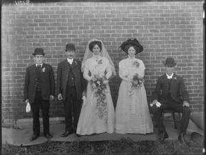 Wedding portrait of unidentified bride and groom with wedding party, against a brick wall background, possibly Christchurch district
