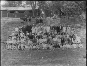 A group of unidentified people, men, women and children sitting on bank, below a shed in an unidentified park, possibly Christchurch district