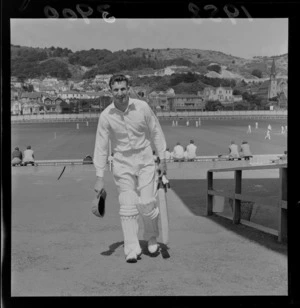 Unidentified cricketer wearing pads and holding cricket bat with game of cricket being played in the background, Basin Reserve, Wellington