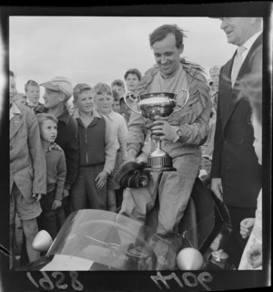 Merv Neil, winner of trophy cup, in his single seater racing car, surrounded by child spectators, Levin motor racing circuit, Horowhenua District