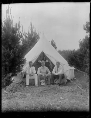 Three unidentified young men sitting outside a tent, one holding a frying pan in which sausages are cooking, in bush, probably Christchurch district