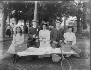 Group of unidentified men, women and girl having a picnic outdoors, probably Christchurch, includes a tennis racquet leaning on table