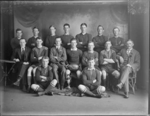 Studio portrait of the Hornby League Football President rugby team for 1919, coaches with rosettes, ball and cup with kewpie doll in front, Christchurch