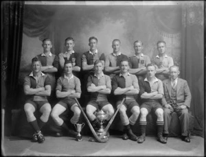 Studio portrait of unidentified men's hockey team in uniforms and coach, with pads, hockey sticks, small and large trophy cup in front, Christchurch
