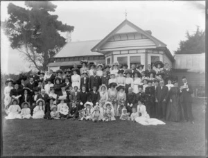 Wedding party portrait on grass in front of wooden house with veranda, unidentified bride and groom with extended family, children in front including flower girls [twins?], women with hats, probably Christchurch region