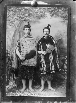 Studio portrait of two unidentified European men in Maori costume, including traditional cloaks and both holding carved weapons [kotiates?], probably Christchurch district
