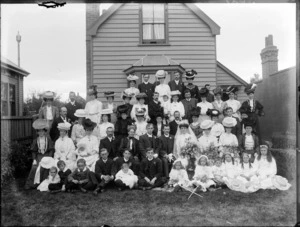 Wedding group portrait in backyard of wooden house, unidentified bride with long veil and groom with extended family, women with hats, children in front, two flower girls with small shepherd's crooks and lace bonnets, probably Christchurch region
