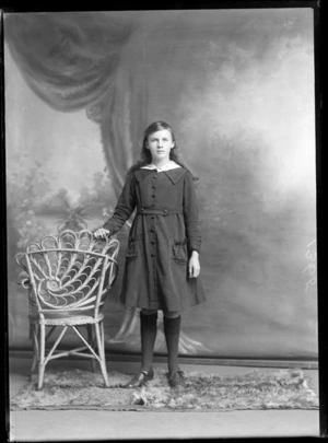 Studio portrait of an unidentified girl, wearing a knee-length dress and socks, beside a wicker chair, possibly Christchurch