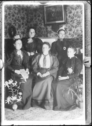 Group of unidentified women indoors, photographs on the wall behind and a vase and pot plant on the floor, probably Christchurch district