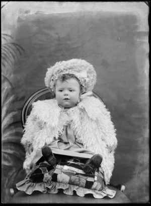Studio portrait of unidentified infant wearing a fur cloak and hat, sitting on a patchwork cushion and chair, probably Christchurch district
