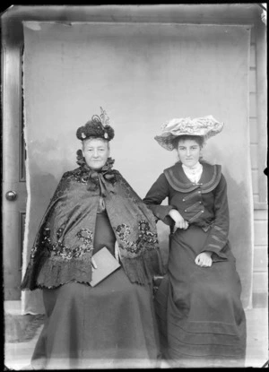 Two unidentified women outside a house, including elderly woman wearing a Victorian sequined cape over dress and both women wearing hats, probably Christchurch district