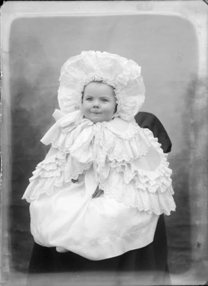 Studio portrait of unidentified baby seated on a covered chair, wearing white lace capelet and bonnet, probably Christchurch district