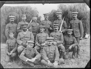 Group photo of soldiers with flags or pennants and dog, while at a social outing, willow trees behind, Hawke's Bay District