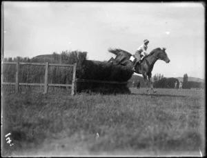 View of a woman and horse jumping a thatched fence in an unidentified competition, Hawke's Bay District