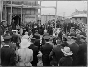 Group, including men in uniform, gathered outside an unidentified building with scaffolding, listening to a speech, Hastings