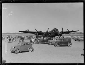 Avro Lancaster aeroplane, at an unidentified aerodrome, surrounded by crowd of men, women and children, and parked motorcars