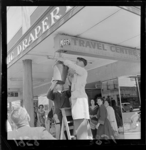 Unidentified men remove birds nest from sign reading 'Air Travel Centre, Rail & Hotel Bookings', Lower Hutt, Wellington Region, with onlooking pedestrians