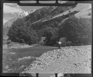 Lake Ohau, Central Otago, view of upper Temple Valley with rock covered riverbed, with man fishing in river, trees on scree slope and snow covered mountains beyond