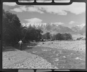 Lake Ohau, Central Otago, view of upper Temple Valley with rock covered riverbed, with man fishing in river by trees, snow covered mountains beyond