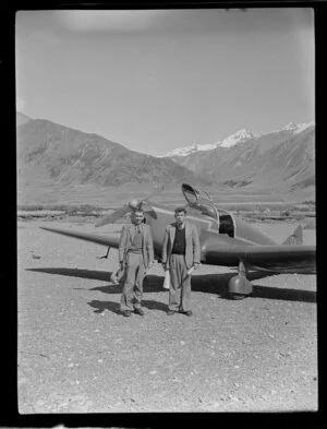 Lake Ohau, Central Otago, view of shingle river bed with two men (one being Leo White) standing beside a Mount Cook Whitley Straight (ZK AUK) tourist aircraft, snow capped mountains beyond