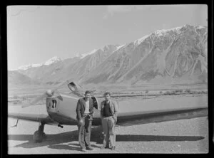 Lake Ohau, Central Otago, view of shingle river bed with two men (one with camera) standing beside a Mount Cook Whitley Straight (ZK AUK) tourist aircraft, snow capped mountains beyond