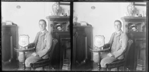 Interior featuring Edgar Richard Williams seated at a table with an award (a shield embossed with a ship) [Williams' Royal Terrace house, Kew?] Dunedin, Otago Region