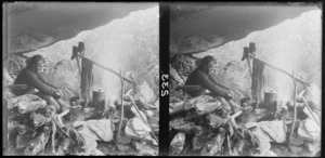Unidentified man cooking and drying his clothes by a fire under an outcrop while tramping [along the Routeburn Track?]