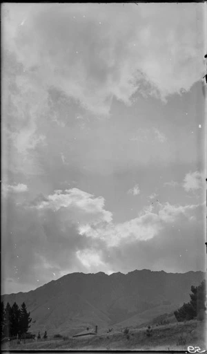 View of a grassy slope, including the roof of a house, and clouds over a mountain in the distance, Queenstown