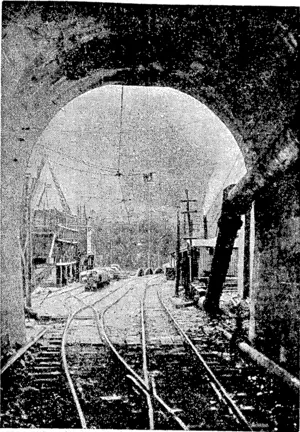 Photograph taken while the tunnel was being driven inside Its Otira end, looking out through West Coast portal, showinfl floor, rails, and wall ef tunnel; foul air exhaust pipe (the'upper one); compressed air main (the lower one); and outside—various structures and the top of the superstructure of the Rolleston River.bridge. – (Evening Post, 04 August 1923)