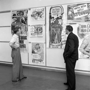Two men looking at a group of posters on a wall advertising movies