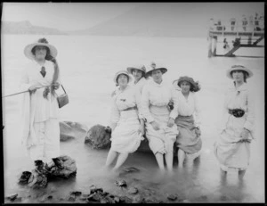 Group of unidentified women, paddling in the shallows [of a lake? sea inlet?], with people on a pier behind, probably Christchurch district