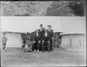 Four unidentified smartly dressed men, sitting with a cockatoo in a birdcage, beside tents, including a sign, which reads 'Flora Camp, 1901-02', and a boy at left of image, location unidentified