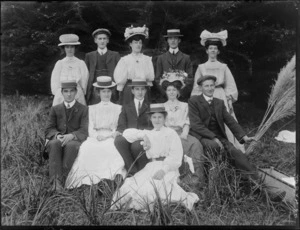 Unidentified group of young women and men, wearing picture hats and straw boaters, some with a small rosette pinned to chest, and one member of party holding a bunch of Toi Toi flowers, in an outdoor location, probably Christchurch district