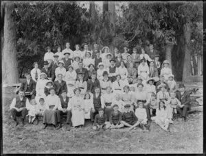 Large group portrait of unidentified men, women and children, sitting outside beneath trees, probably Christchurch district