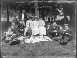 Unidentified group picnicking, showing women, men, and boys sitting around food which is laid out on a cloth, beneath a tree, whose trunk has been carved with peoples' names