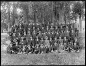 Unidentified group of men, all wearing rosettes attached to their lapels, and some hold brass instruments, posed among a stand of eucalyptus trees, probably Christchurch district