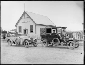 Two motorcars, one of which belongs to Smiths Taxicabs Ltd, with passenger and drivers, on road in front of a wooden hall, which has a sign reading 'Orange Hall' on front, probably Christchurch district